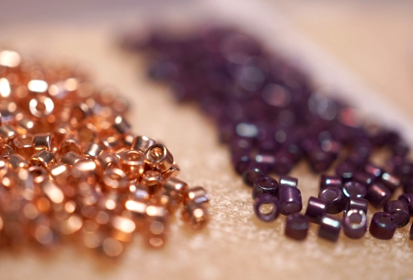 MIYUKI Delica beads - photo by Crafty Bead A macro close-up of two piles of shimmering Miyuki Delica beads. The foreground shows shiny copper-colored cylindrical beads, while the background pile is a deep purple. The photo highlights their perfect shape for peyote stitch.